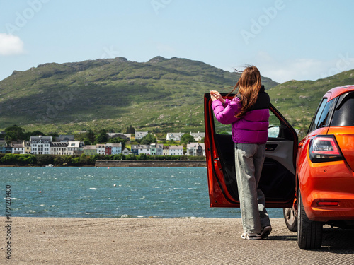 Young girl is standing by a small red sport car and looking at stunning nature scenery with ocean, Roundstone town and Errisbeg Mountain in the background. County Galway, Ireland. Travel and tourism