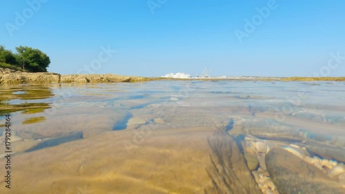 Calm water gently flowing over rocks on sunny day. Crystal-clear water gently washes over submerged rocks near the shore, creating a tranquil scene on a bright, sunny day, offering a serene view