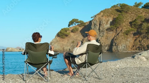 Couple relaxing on beach chairs, enjoying coffee and scenic view. Tourists enjoy a relaxing moment on a pebble beach, sipping coffee from mugs while admiring the breathtaking view of a tranquil bay
