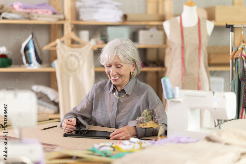 Fototapeta premium Positive old tailoress drawing dress design in tablet while sitting at table in sewing workshop