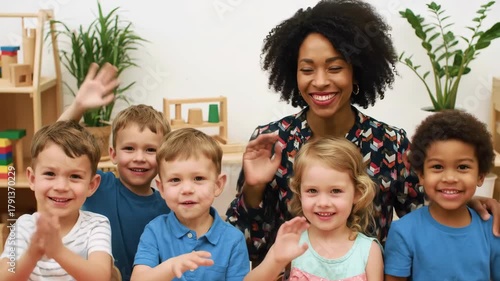 Cheerful scene of a female teacher with curly afro and diverse group of young children joyfully waving hands in a sunlit classroom with play blocks and wooden shelves