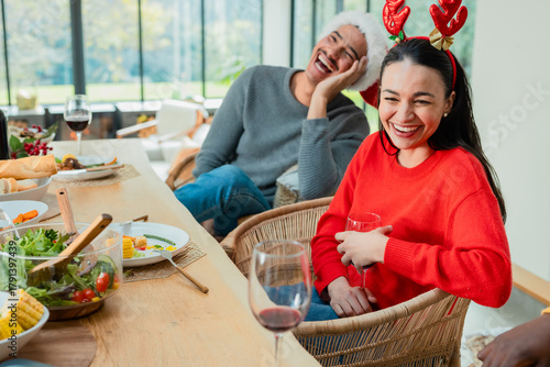 Couple laughing wearing sweaters holding wine glasses at dining table near windows with corn on cob