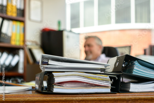 A desk with piles of paperwork in the foreground and a smiling worker, blurred in the background - horizontal