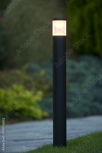 A sleek, black LED bollard light illuminates a garden path and lawn at dusk, with a soft, warm glow emanating from its angular geometric top, set against a blurred green background.