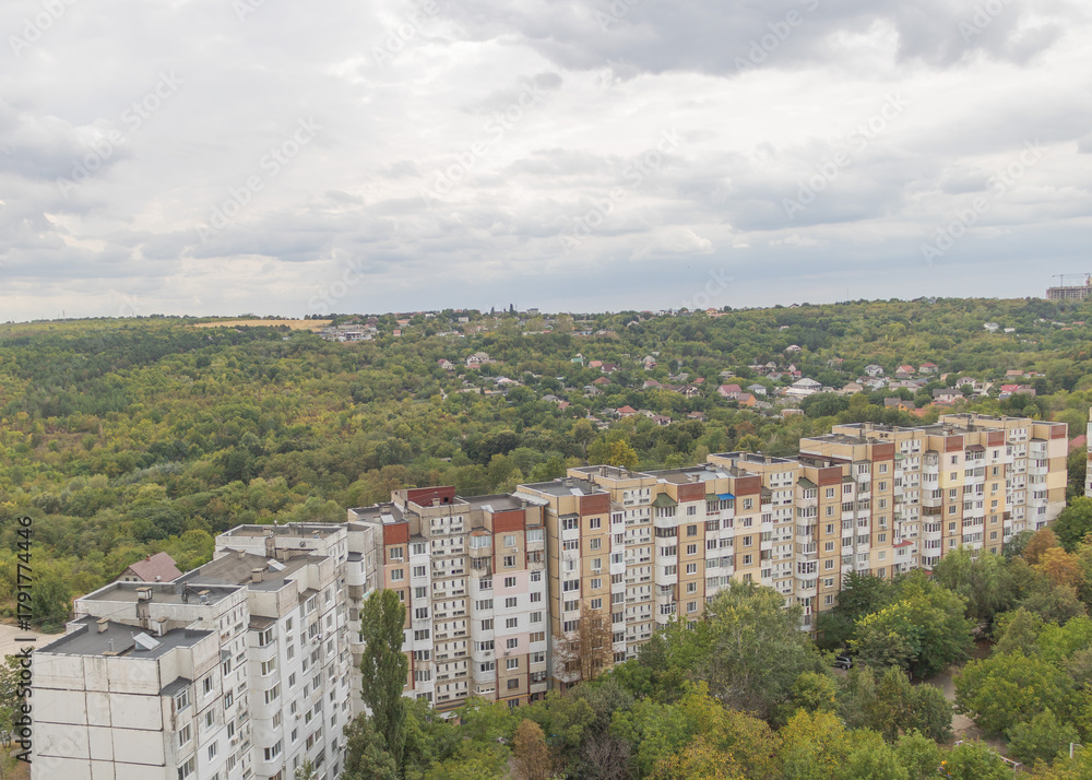 Fototapeta premium Aerial view of residential apartment blocks near lush green forest and suburban hillside under cloudy sky