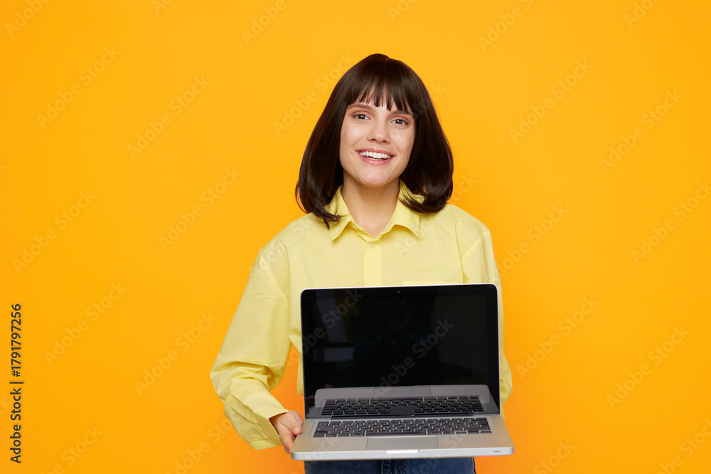Naklejka premium A cheerful woman stands against a bright orange backdrop, holding an open laptop with a friendly smile, conveying modern work life, remote learning, and digital creativity.