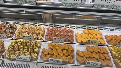 Various types of sushi are displayed on a retail shelf behind a clear glass window. Customers can view and select the fresh food, which is priced and protected for hygiene and quality.
