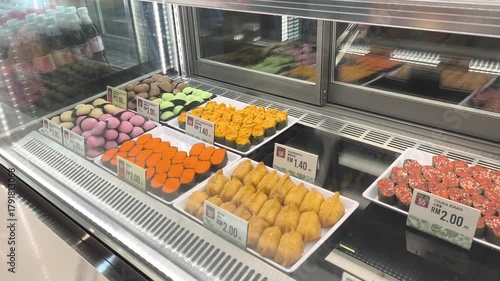 Various types of sushi are displayed on a retail shelf behind a clear glass window. Customers can view and select the fresh food, which is priced and protected for hygiene and quality.
