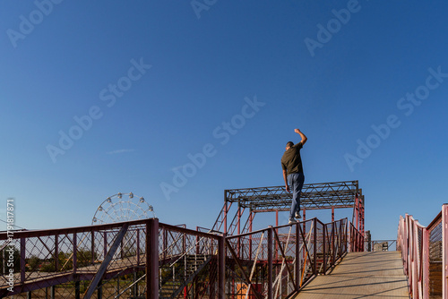 Freerunner walks across high railing in outdoor parkour structure, raising arm for balance against clear blue sky with Ferris wheel in distance. Precision in urban acrobatic movement. Parkour performa