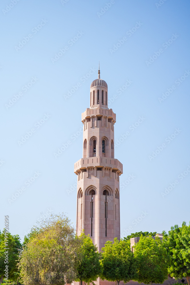 Fototapeta premium A tall Islamic minaret tower rising above lush green trees under a bright, cloudless blue sky in Oman. The tower features elegant arches, beige stonework, and traditional Middle Eastern architecture