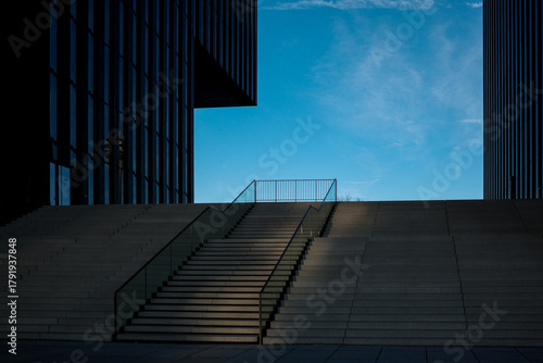 A wide outdoor staircase with glass railings leads upward between two modern commercial buildings with dark slatted facades.