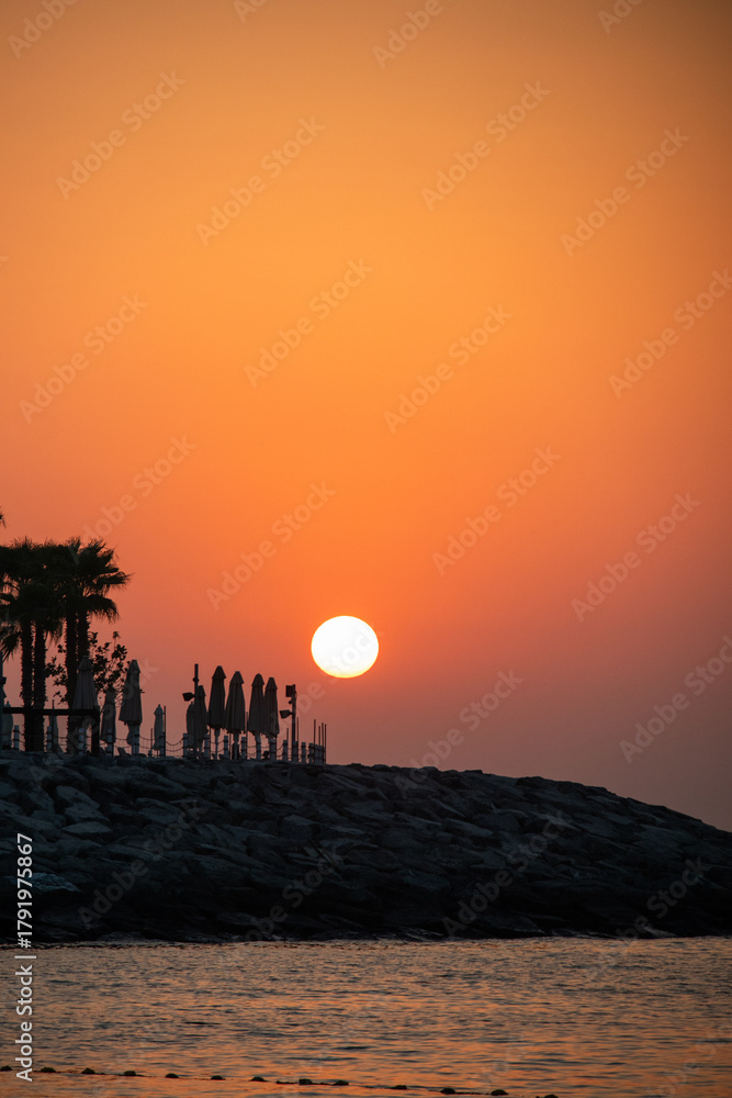 Fototapeta premium A dramatic, intense orange and red sunset or sunrise over the sea, silhouetting beach umbrellas and palm trees on a rocky shoreline