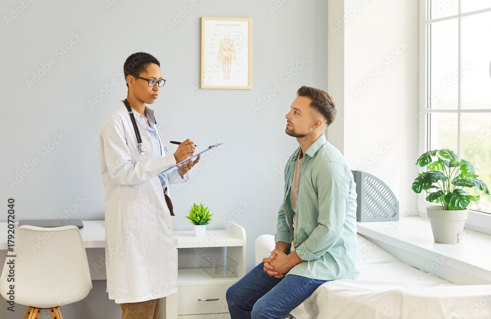 Fototapeta premium Portrait of female doctor talking with young male patient sitting on couch in clinic in exam room. Physician giving consultation to a man during medical examination in office. Medicine concept.