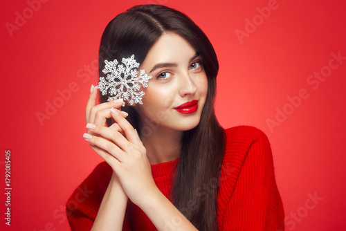 Beautiful young woman with red lipstick, shiny clean skin and face, showing glowing snowflake, near face and smiling, isolated on a red background. Christmas holidays concept