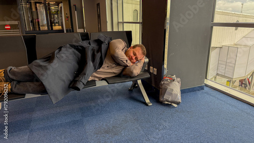 A weary traveler, Caucasian male, dozes on an airport bench, embodying the spontaneous serenity of Lost Luggage Day