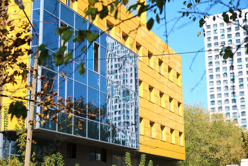 Photos Modern office building with bright yellow facade panels and mirrored glass windows reflecting a nearby high-rise tower