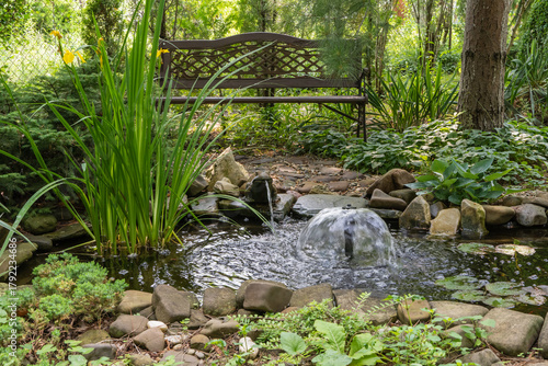 Papier peint Serene garden pond with small waterfall, surrounded by rocks, lush greenery, and tall plants