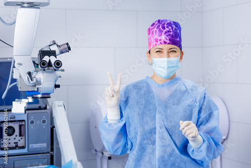 Foto Ophthalmic surgeon with mask showing peace sign beside operating microscope