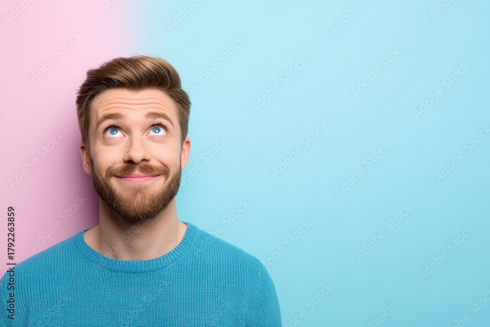 Fototapeta premium A man with a beard looks upwards, wearing a blue sweater, against a pink and blue background.