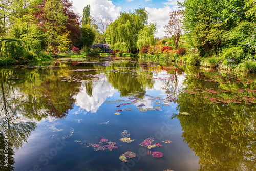 Peaceful garden scene with water lilies, vibrant greenery and cloud reflections on a calm pond surface in Giverny, France.