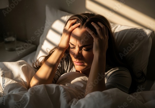 Woman in bed holding her head in pain, illuminated by sunlight streaming through blinds