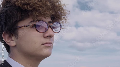 Portrait of a curly-haired teenager with glasses and a mature business suit against a backdrop of a hill and blue sky. The camera pans around.