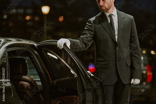 Caucasian middle aged man wearing suit and gloves opening car door outdoors at night, standing beside luxury vehicle, performing professional chauffeur service