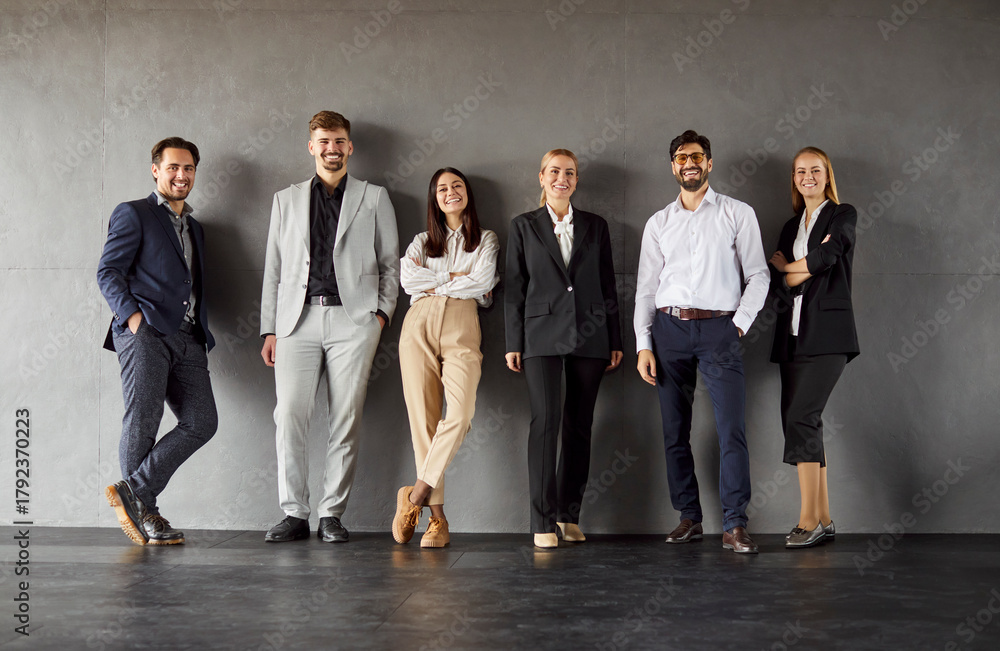 Obraz premium Group full length portrait of confident business professionals standing against plain wall, smiling and looking at camera. Diverse team in formal and smart casual attire, showing unity and positivity.