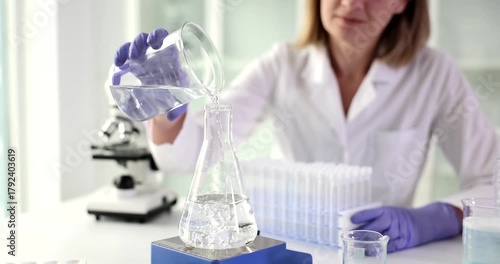 Woman laboratory assistant in white coat pours water into flask standing on heater. Laboratory worker carefully observes reaction with full attention