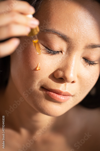 Wallpaper Mural Asian woman applying facial serum with dropper, showcasing skincare routine, illuminated by soft natural light, emphasizing beauty and self-care practices for healthy skin Torontodigital.ca