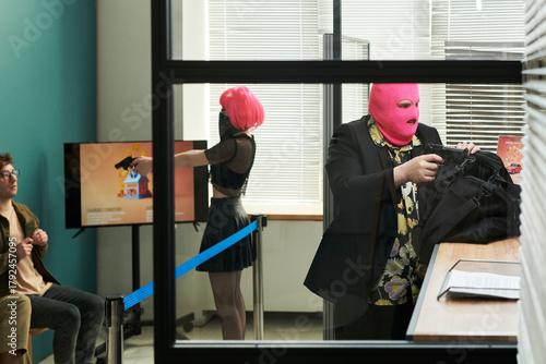 Two masked young adults, one Caucasian man wearing pink balaclava putting bag on counter, one woman with pink wig pointing handgun at seated Caucasian man in modern bank setting