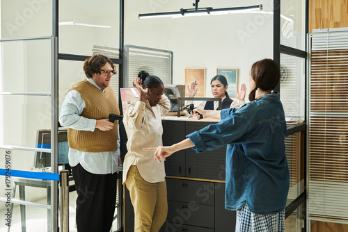 Caucasian man and woman holding guns confronting Black woman and Hispanic woman with hands raised in bank office setting, displaying tense expressions during standoff