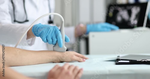 Patient arm rests on table while ultrasound probe touches wrist for scan. Clinician scans tendons and veins moving transducer to record findings