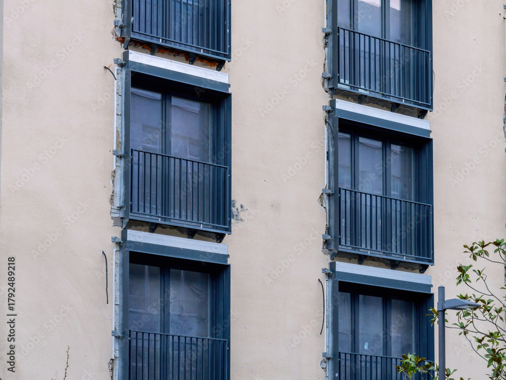 Obraz premium Facade of a building under reconstruction showing multiple blue-framed windows with small metal balconies, exposed edges, and visible structural reinforcements against a beige wall
