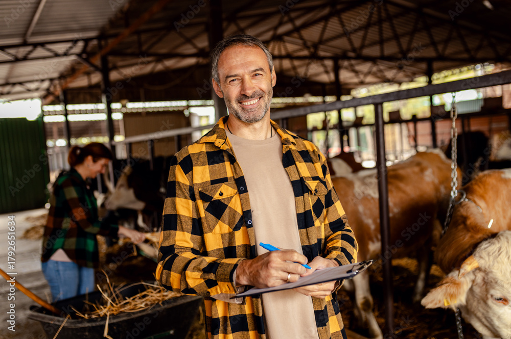 Fototapeta premium Male farmer managing a cattle barn with a female worker, making notes on clipboard.