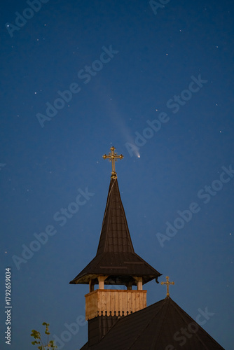 Celestial and Spiritual: Rare Comet ATLAS 2024 Shines Brightly in the Night Sky Behind the Illuminated Cross of an Old Wooden Church, Symbolizing Faith and Science