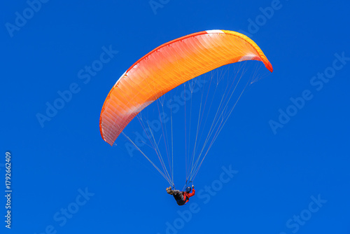 A single paraglider with a vibrant orange and red canopy is seen against a deep, clear blue sky during a sunny day flight.