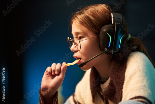 Fotografie Caucasian teenage girl wearing headset eating potato chip while gaming on comput
