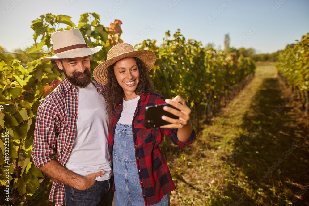 Fototapeta premium A couple stands among lush vines in a vineyard, smiling as they take a selfie on a sunny day. Their joy reflects their passion for family wine production and shared experiences.