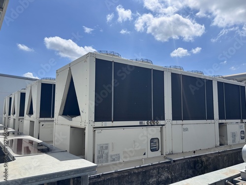 A row of large, modular commercial air-cooled chillers and HVAC units sits on a rooftop under a bright, partly cloudy blue sky. These systems are essential for industrial climate control and building 