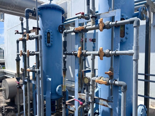 Two tall, blue industrial water filtration tanks are connected by a complex system of piping, valves, and gauges against a plain white wall. This scene illustrates the infrastructure of a commercial 