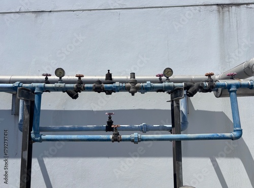 Industrial pipework system on an exterior building wall, featuring a backflow preventer assembly, gate valves, and pressure gauges on galvanized and blue-painted water pipes under bright sunlight.