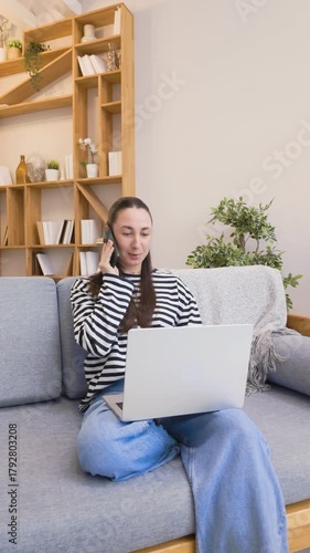 Young woman talks on smartphone while sitting on couch with laptop at home, ending call and placing phone beside her