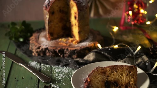 Two slices of traditional panettone are presented on small white plates on a dark green wooden table dusted with powdered sugar, set against a backdrop of warm holiday string lights and a whole loaf. 