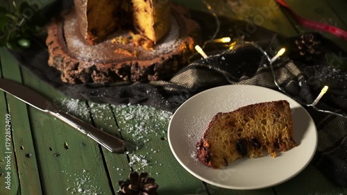 A single slice of moist panettone, dusted with powdered sugar, is the focus of a rustic holiday scene on a deep green wooden table, surrounded by pine cones and twinkling string lights. The whole pane