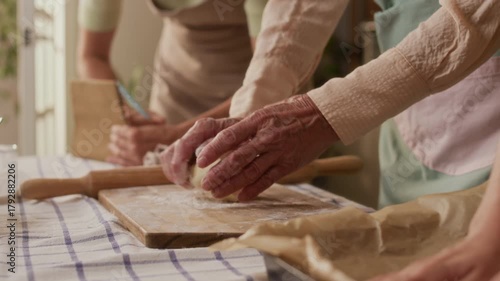Close up of grandmothers hands kneading dough on wooden board teaching adult daughter and granddaughter homemade puff pastry recipe while cooking together in kitchen