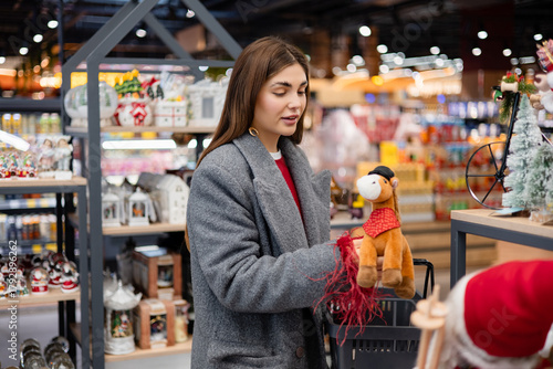 Young woman placing a Christmas toy into a shopping basket while browsing festive decorations in a holiday store.