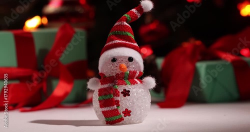 A snowman in a colorful hat stands cheerfully among wrapped gifts, decorated for Christmas. The background is filled with warm holiday lights, creating a joyful atmosphere for the New Year.