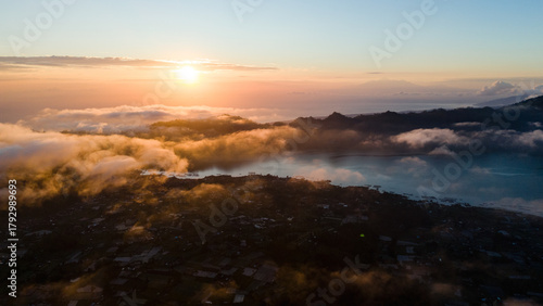 Aerial view of the sun kissing the clouds over the rugged terrain, painting the sky with hues of orange and gold, Mount Batur, Bali, Indonesia.