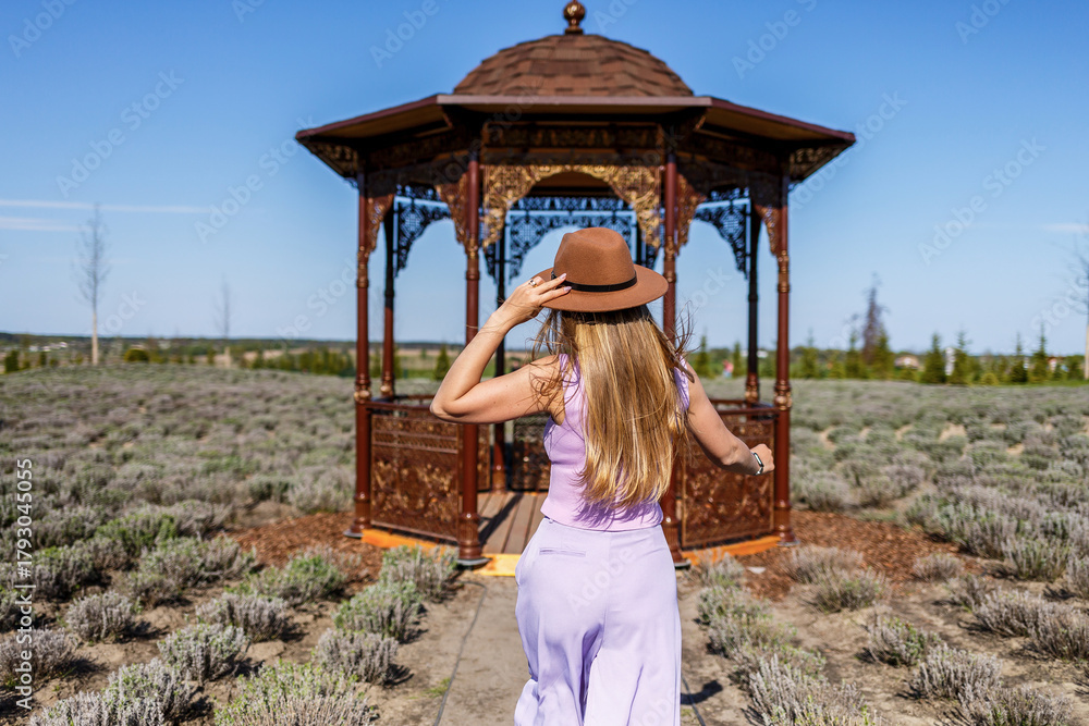 Obraz premium Woman in a purple dress approaches a charming gazebo surrounded by blooming lavender fields under a clear blue sky